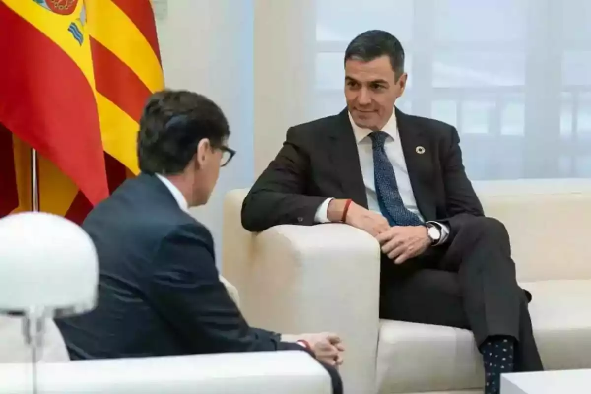 Dos homes amb vestits foscos conversen asseguts en un sofà blanc amb banderes de fons.