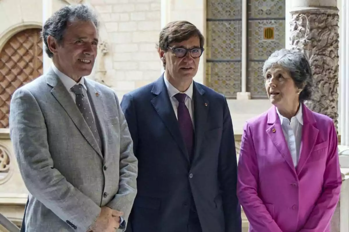 Tres persones dretes en un entorn arquitectònic elegant, amb vestits formals i expressions serioses.
