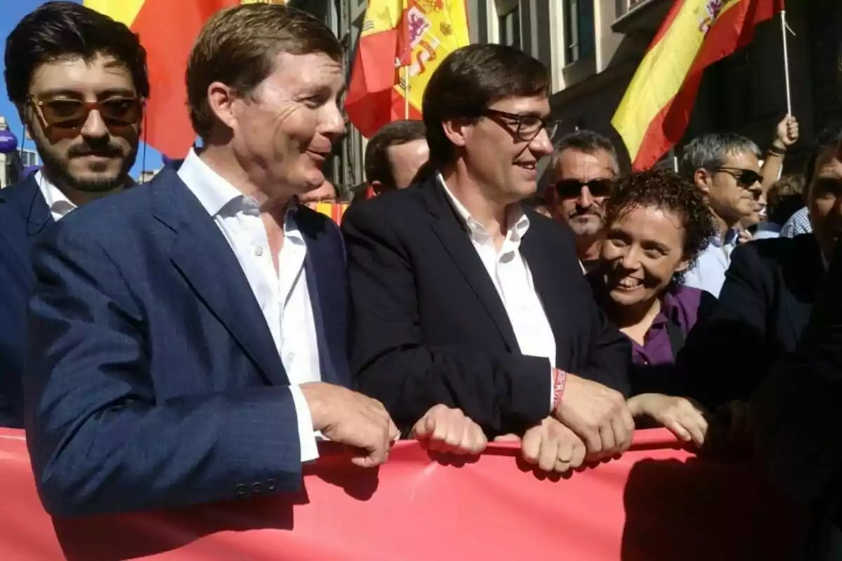 Un grup de persones somrients sosté una pancarta vermella en una manifestació amb banderes espanyoles de fons. Un grup de persones somrients sosté una pancarta vermella en una manifestació amb banderes espanyoles de fons.