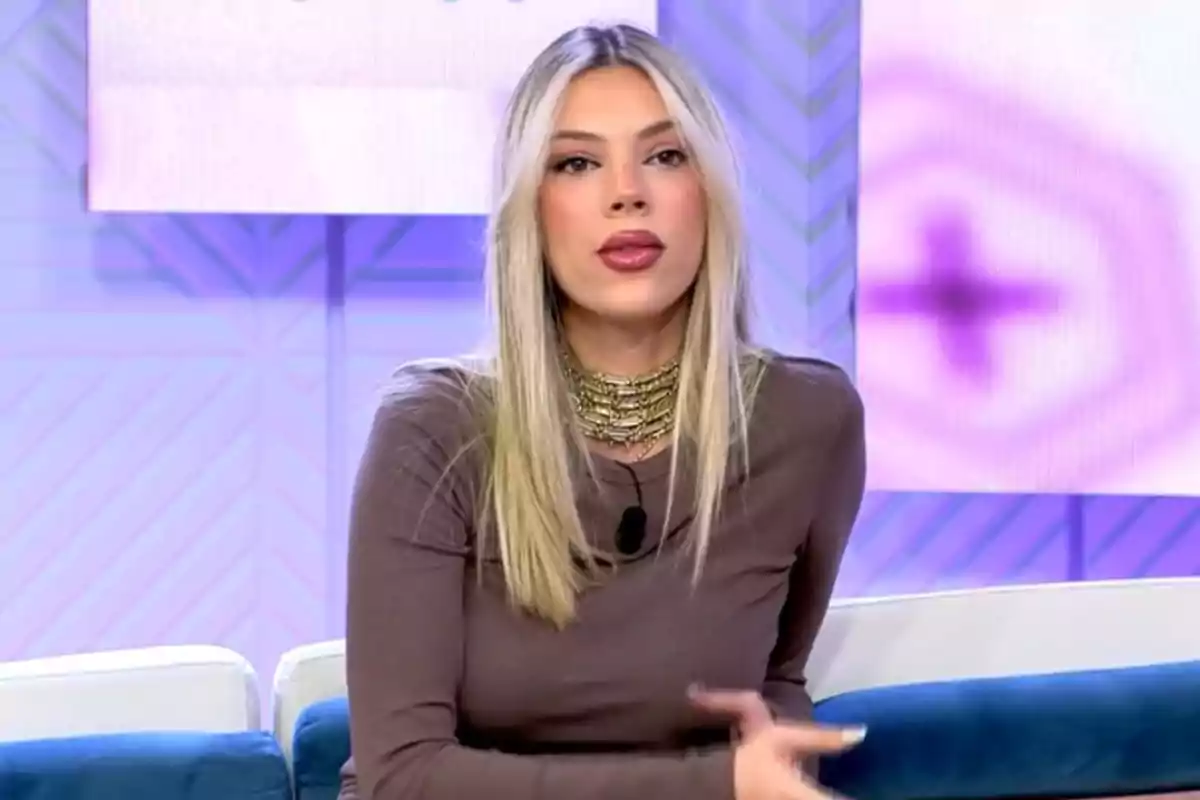 Una dona amb cabell ros i llarg està asseguda en un sofà durant un programa de televisió, porta un vestit marró i un collaret daurat, el fons és de tons morats i blaus. Una dona amb cabell ros i llarg està asseguda en un sofà durant un programa de televisió, porta un vestit marró i un collaret daurat, el fons és de tons morats i blaus.
