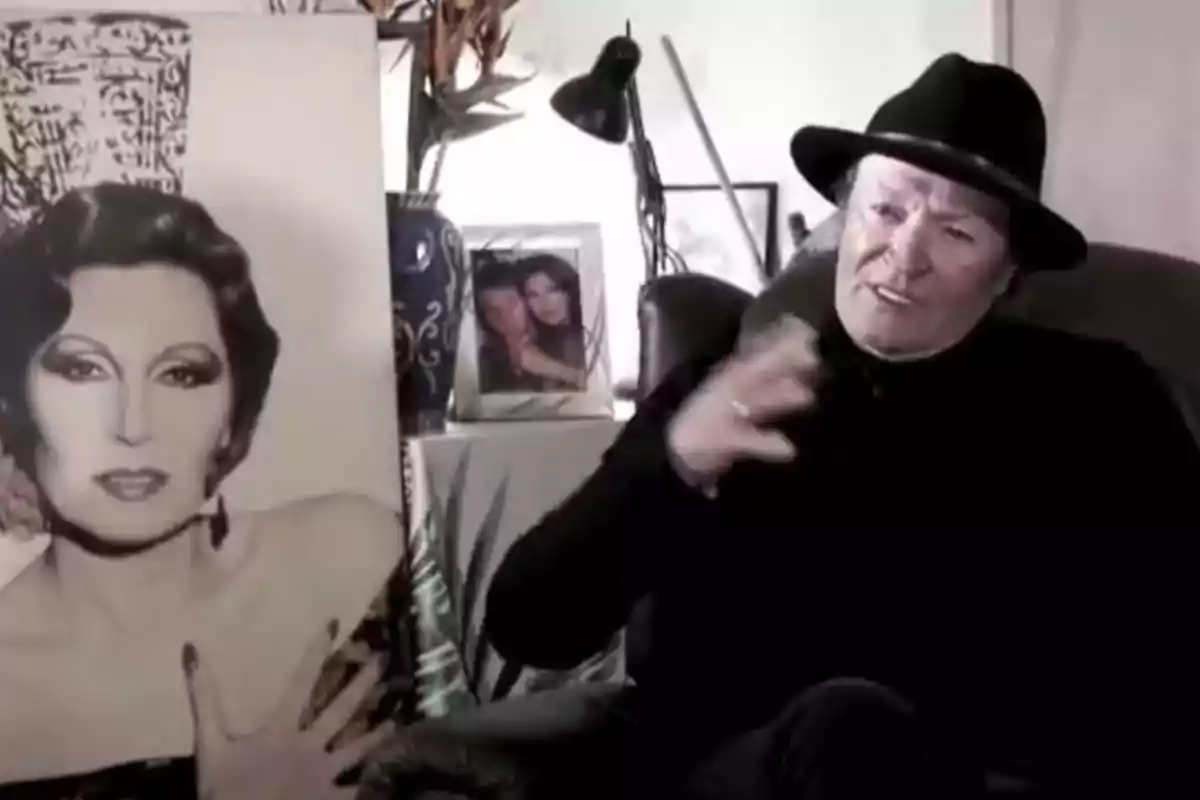 Amador Mohedano with a black hat is sitting in a room next to a portrait, with a framed photograph in the background. Amador Mohedano with a black hat is sitting in a room next to a portrait, with a framed photograph in the background.