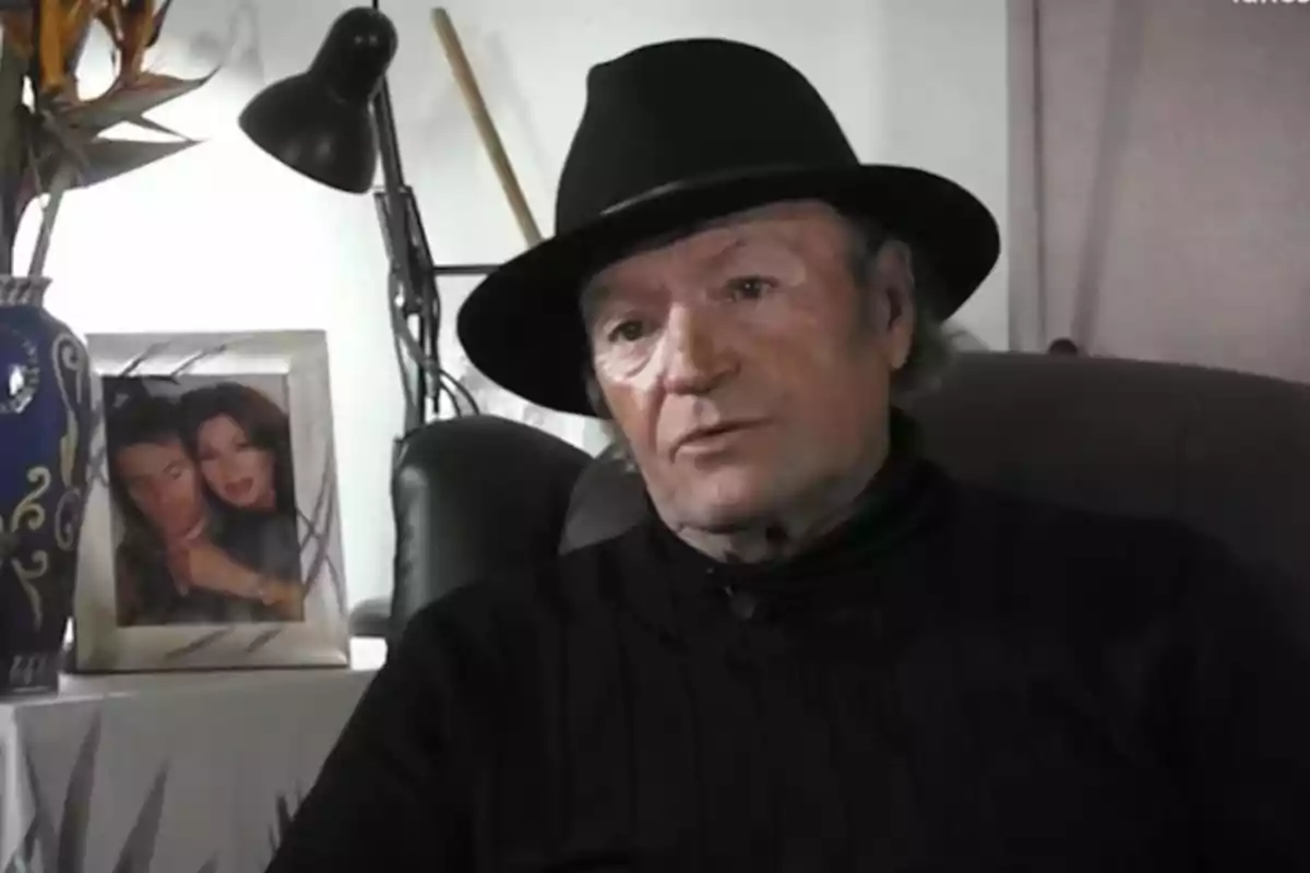 Un home amb barret negre està assegut en un sofà al costat d'una taula que té un gerro decoratiu i un marc de fotos amb una foto de dues persones. Un home amb barret negre està assegut en un sofà al costat d'una taula que té un gerro decoratiu i un marc de fotos amb una foto de dues persones.