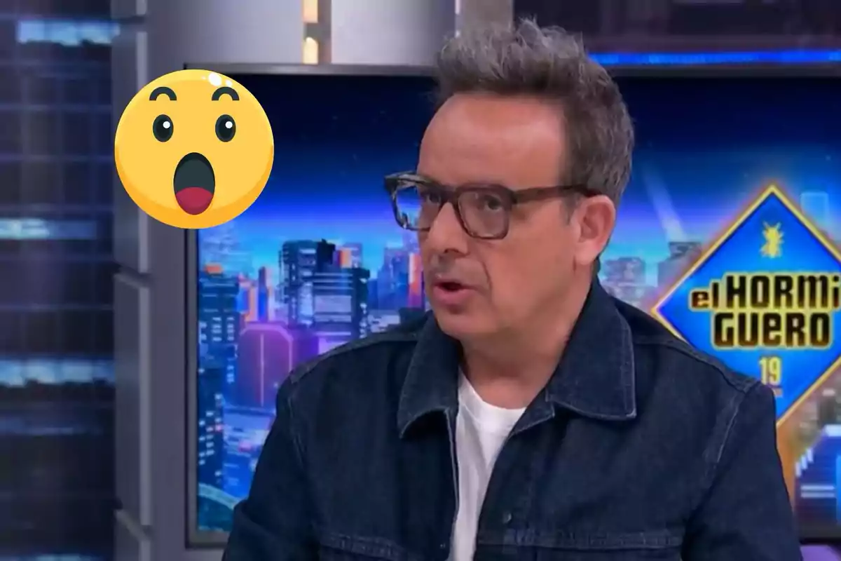 Àngel Llàcer con gafas y chaqueta de mezclilla está hablando en un programa de televisión con un fondo colorido y un emoji sorprendido flotando cerca.