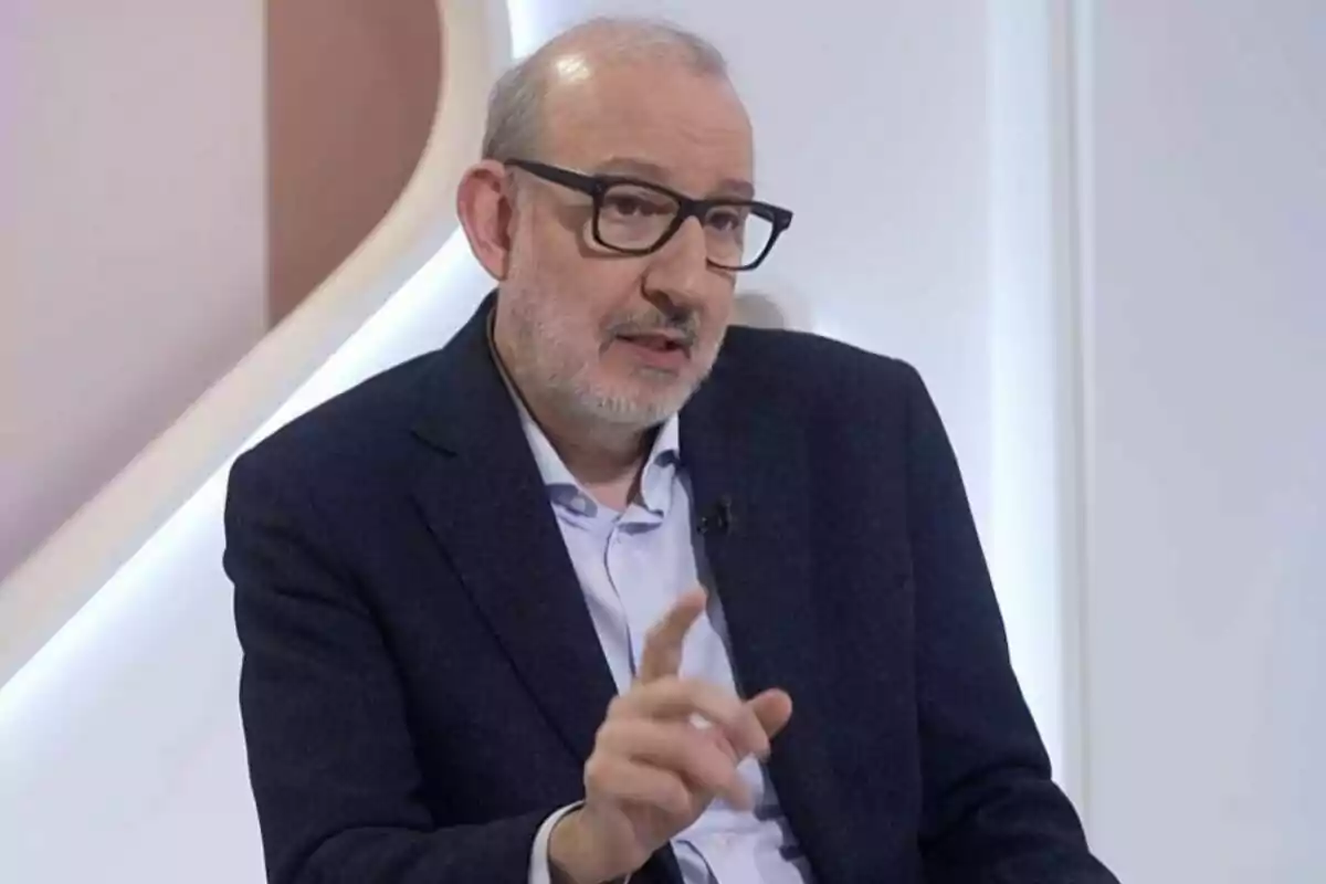 A man with glasses and a graying beard is sitting and gesturing with his hand while speaking in a well-lit environment.