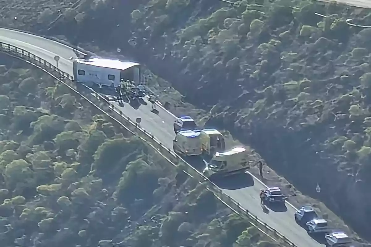 Una carretera de muntanya amb un accident de trànsit on un camió ha bolcat i hi ha diversos vehicles d'emergència a l'escena. Una carretera de muntanya amb un accident de trànsit on un camió ha bolcat i hi ha diversos vehicles d'emergència a l'escena.