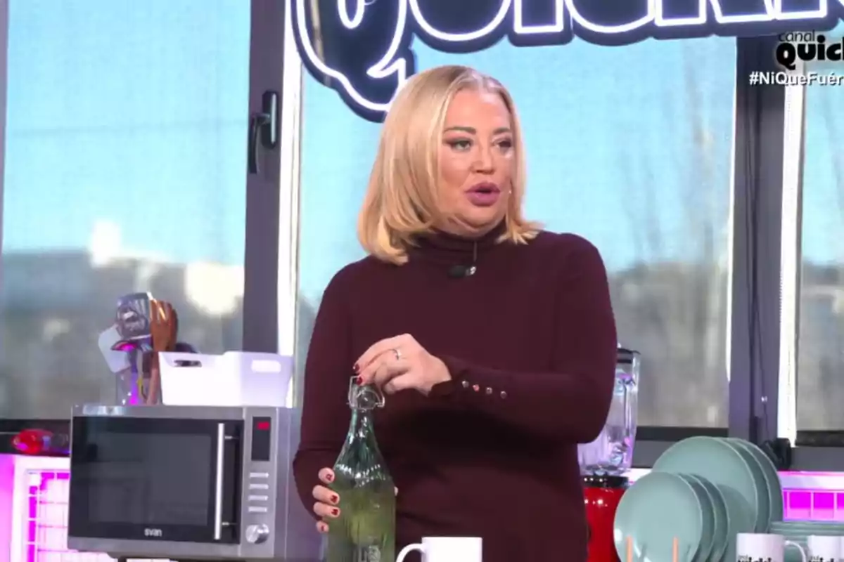 Belén Esteban de cabello rubio en un set de cocina con una botella verde en la mano y utensilios de cocina al fondo.