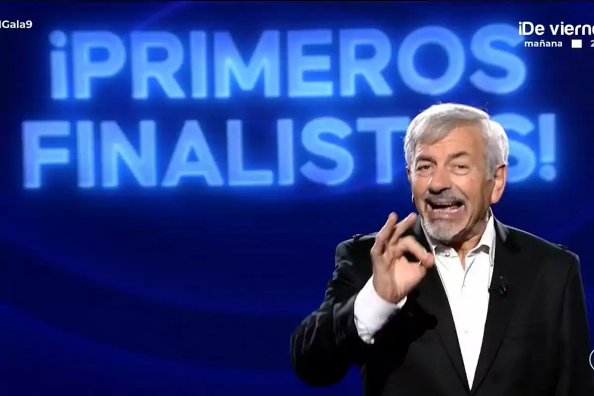 Carlos Sobera de cabello canoso y barba está en un escenario con un fondo azul de 'GH Dúo' que dice "¡PRIMEROS FINALISTAS!" y hace un gesto con la mano.