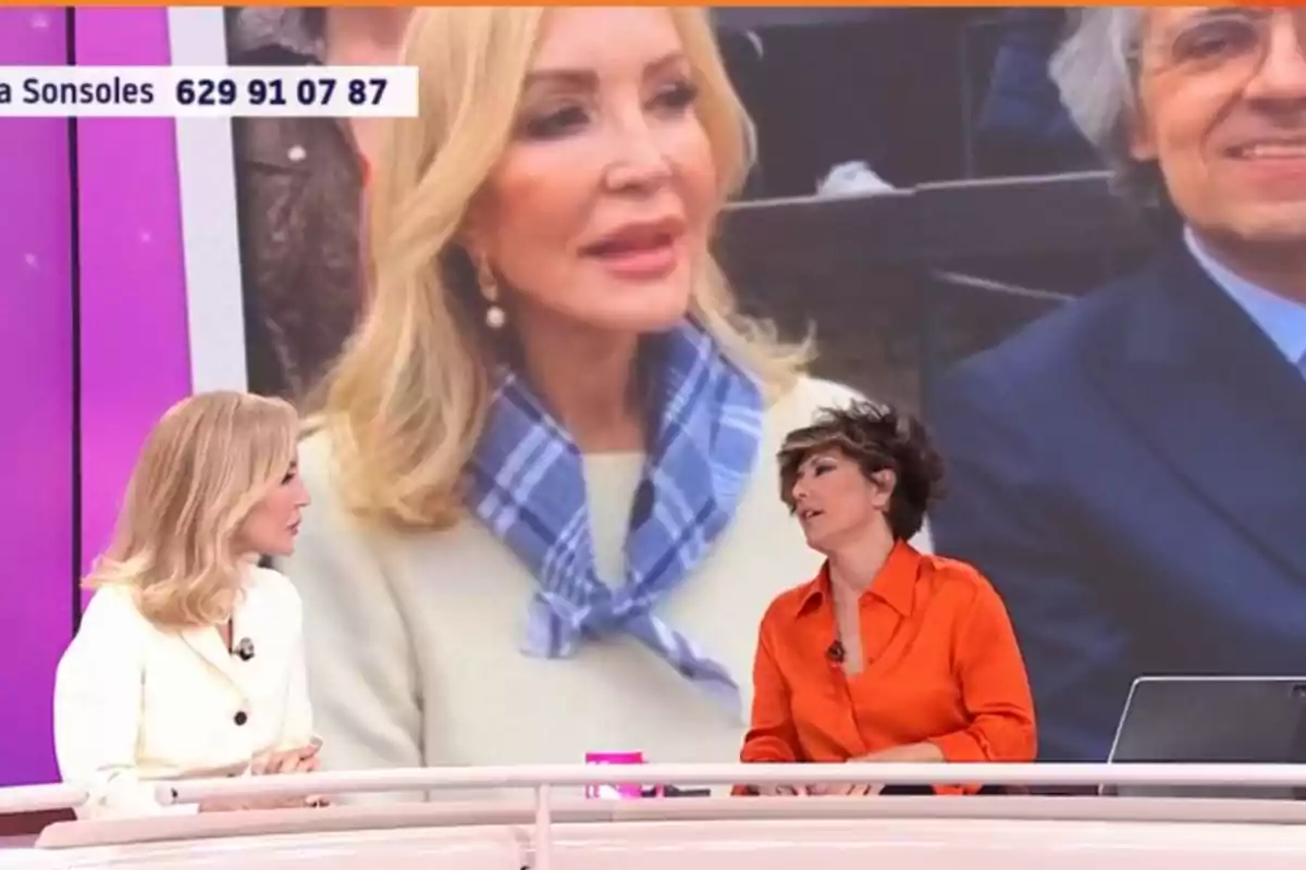 Carmen Lomana y Sonsoles Ónega conversan en un set de televisión, Carmen viste de blanco y Sonsoles de naranja, mientras en el fondo se ve una imagen ampliada de otra mujer con cabello rubio y un pañuelo azul. Carmen Lomana y Sonsoles Ónega conversan en un set de televisión, Carmen viste de blanco y Sonsoles de naranja, mientras en el fondo se ve una imagen ampliada de otra mujer con cabello rubio y un pañuelo azul.