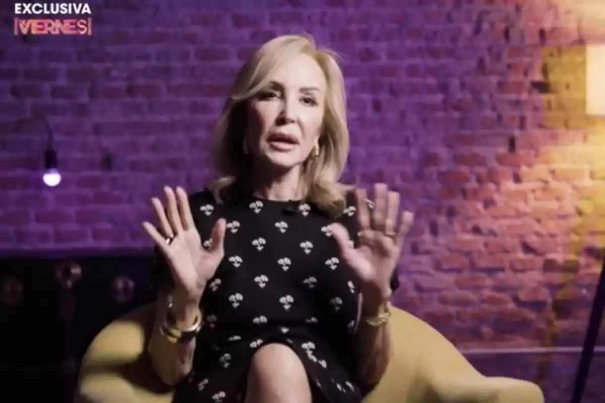 A woman sitting in a yellow chair with a brick background lit in purple tones, gesturing with her hands.