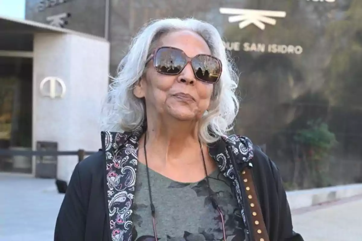 A woman with gray hair and sunglasses is standing in front of a building.