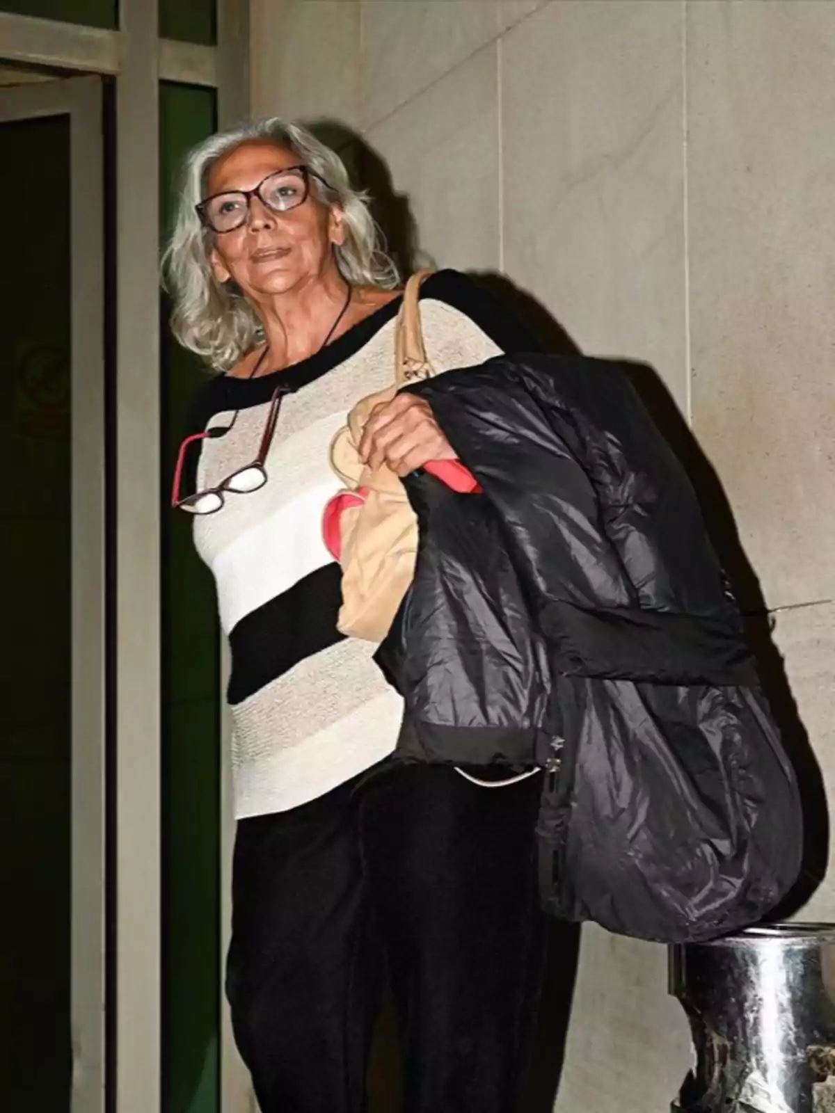 A woman with gray hair and glasses holds a dark coat while standing in front of a door.