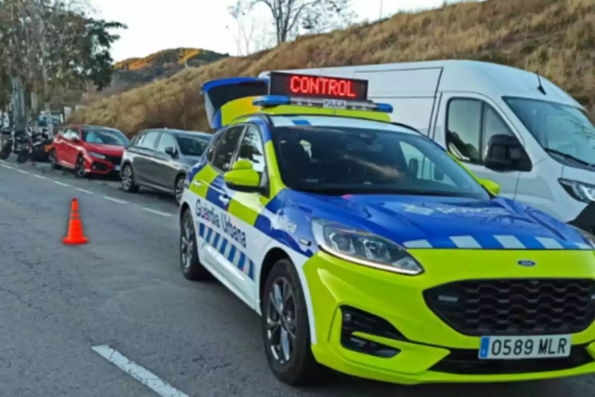 Un cotxe de la Guàrdia Urbana de Barcelona amb un rètol de control al sostre està estacionat en una carretera al costat d'altres vehicles i un con taronja.