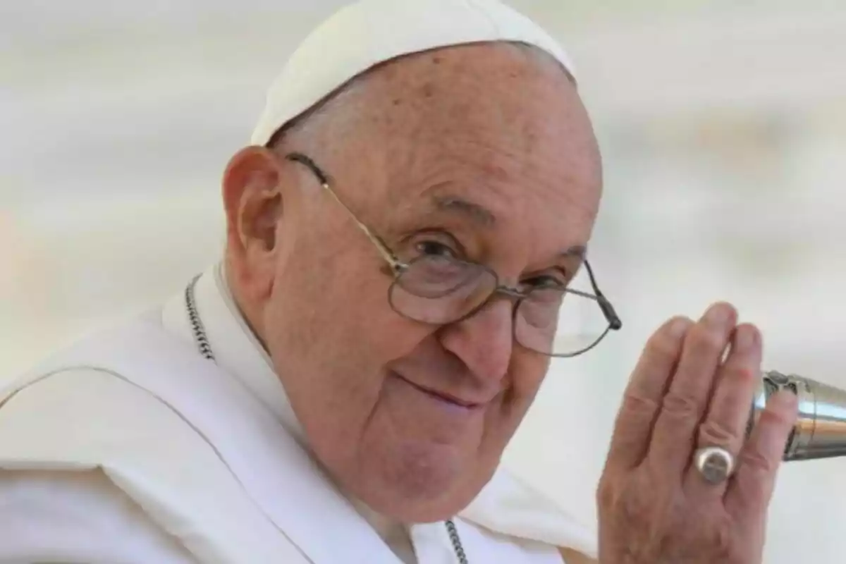 Close-up of Pope Francis with glasses and white attire smiling and raising his hand in greeting.