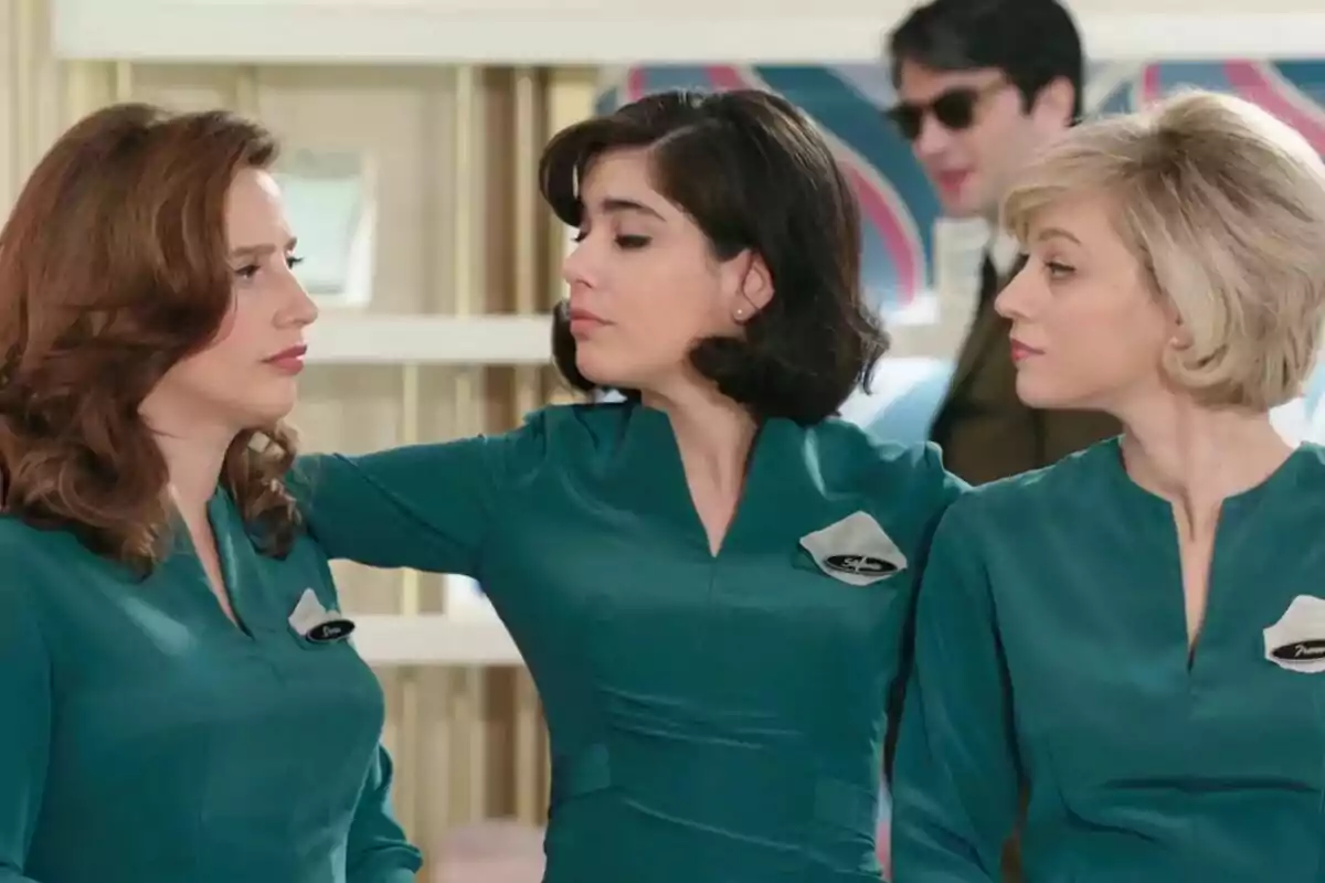 Three women in green uniforms chat as a man wearing sunglasses walks past.