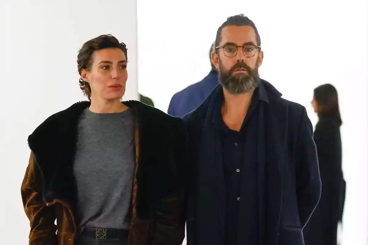 Two people standing, a woman in a brown jacket and a man with a beard and glasses, both looking forward in a well-lit indoor setting. Two people standing, a woman in a brown jacket and a man with a beard and glasses, both looking forward in a well-lit indoor setting.