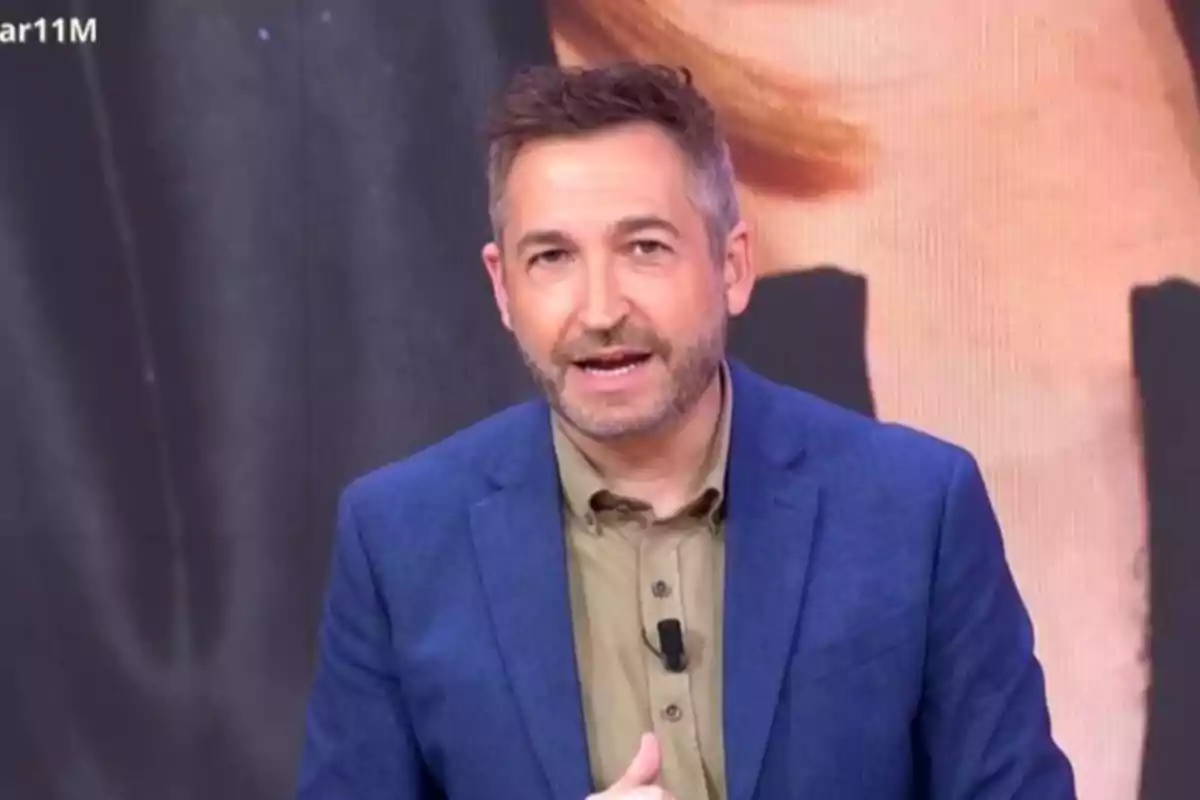 Frank Blanco con barba y cabello corto lleva un traje azul y camisa beige mientras habla frente a una cámara.