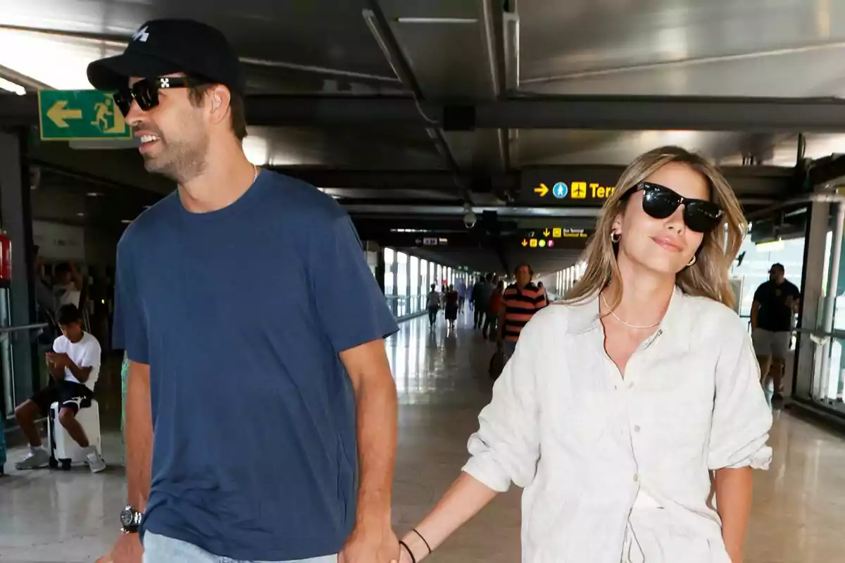 A couple walks hand in hand at an airport, both wearing sunglasses and looking relaxed as they move down the hallway. A couple walks hand in hand at an airport, both wearing sunglasses and looking relaxed as they move down the hallway.