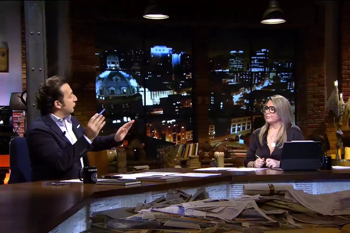 Iker Jiménez y Carmen Porter conversan en un estudio de televisión con una vista nocturna de la ciudad al fondo, rodeados de papeles y libros sobre una mesa.