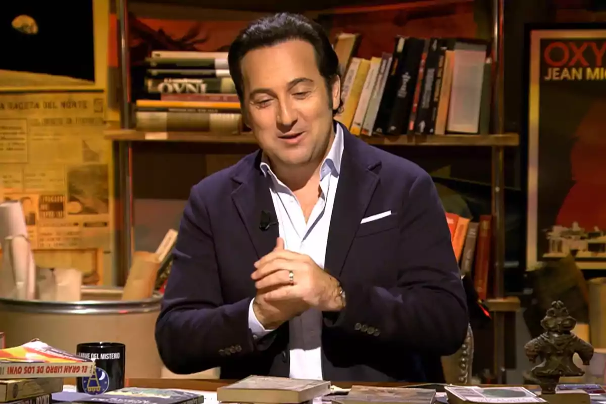 A man in a dark suit and white jersey is sitting at a desk surrounded by books and papers, with a background of bookshelves filled with books and a poster on the wall. A man in a dark suit and white jersey is sitting at a desk surrounded by books and papers, with a background of bookshelves filled with books and a poster on the wall.