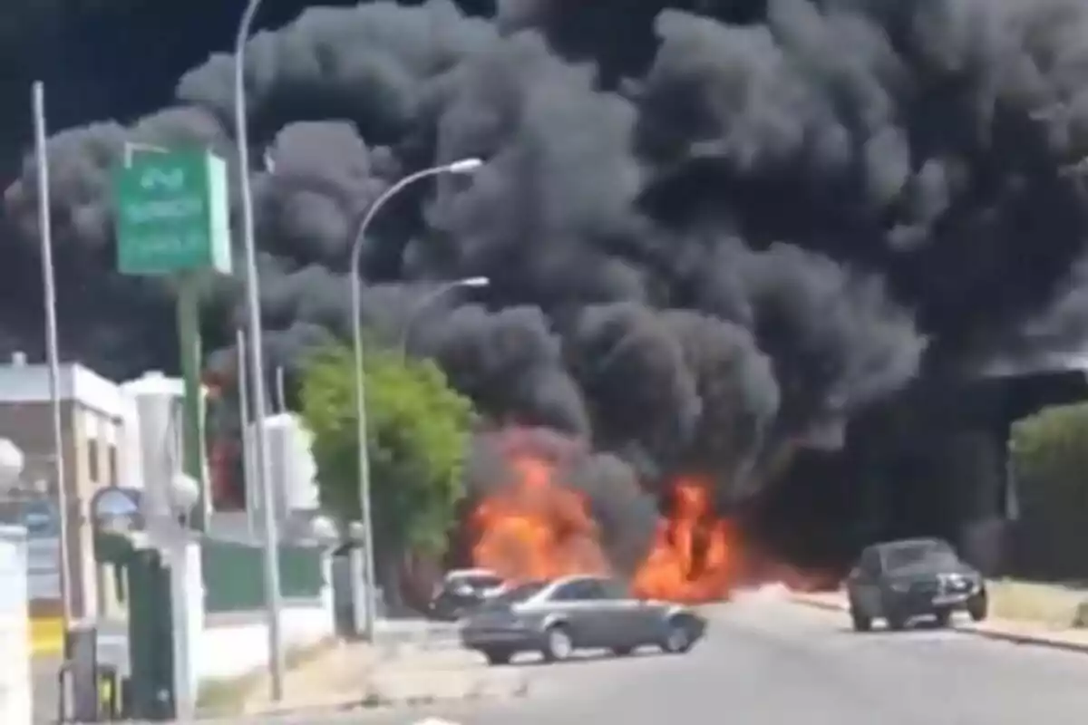 Un gran núvol de fum negre s'eleva al cel mentre les flames cremen intensament en un carrer, amb diversos cotxes estacionats a prop i edificis al fons, a Sevilla.