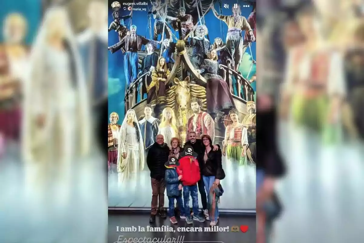 A group of people poses in front of a colorful mural depicting a theatrical scene with characters on a boat. A group of people poses in front of a colorful mural depicting a theatrical scene with characters on a boat.