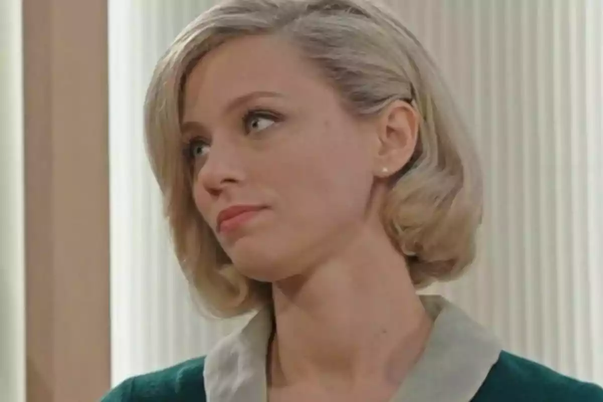 A woman with short blonde hair and a thoughtful expression looks to the side in an indoor setting.