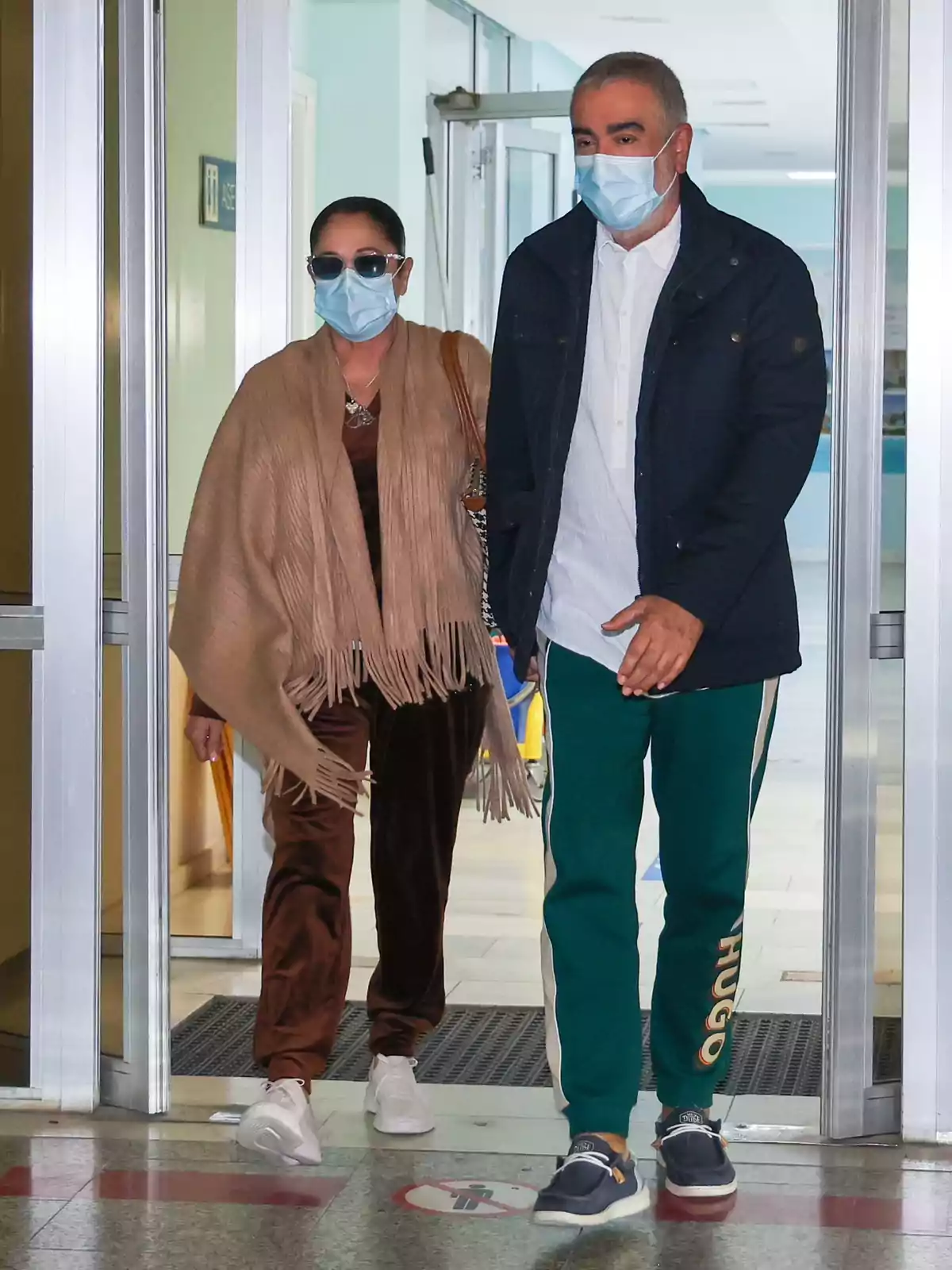 Isabel Pantoja y Agustín Pantoja con mascarillas y gafas de sol salen de un edificio a través de una puerta de cristal.