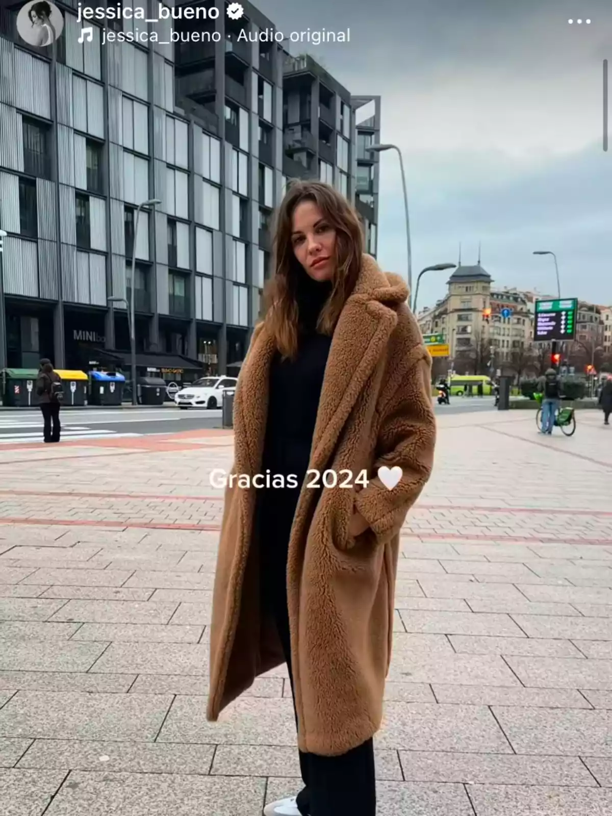 Jessica Bueno con un abrigo marrón posa en una calle urbana con edificios modernos al fondo.