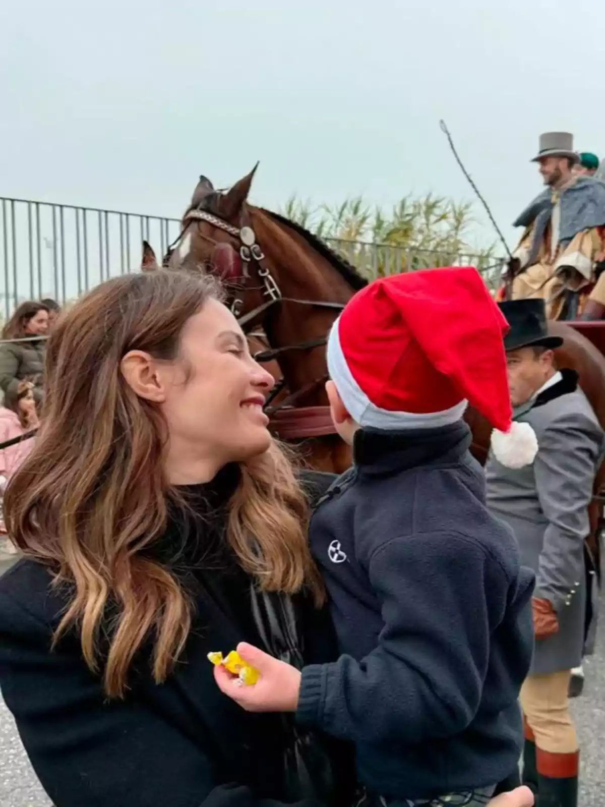 Jessica Bueno sonríe mientras sostiene a un niño con gorro de Navidad, con un caballo y personas vestidas de época al fondo.