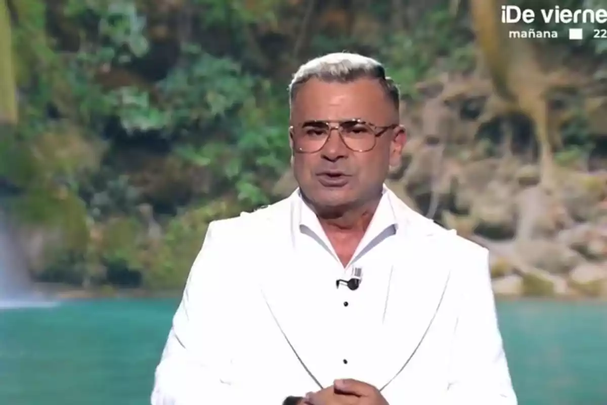 Jorge Javier Vázquez con gafas y cabello canoso está vestido con un traje blanco, de pie frente a un fondo natural con vegetación y agua.