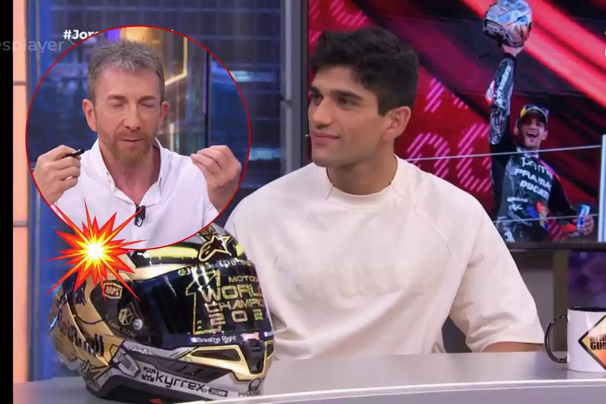 Dos homes en un programa de televisió, un sosté un casc de motocicleta amb la inscripció "World Champion 2023" mentre l'altre sembla estar parlant, al fons es veu una imatge d'un pilot celebrant.