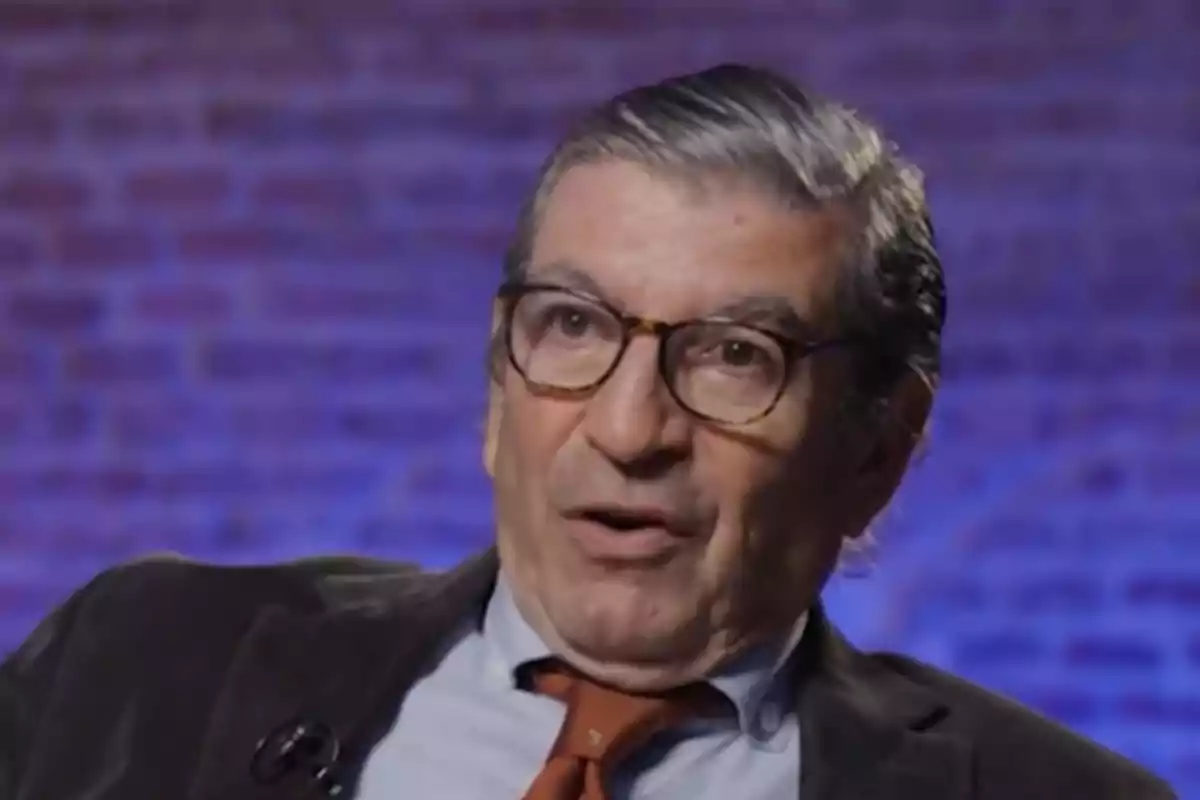 A man with glasses and a tie speaks in front of a brick background lit in blue tones. A man with glasses and a tie speaks in front of a brick background lit in blue tones.