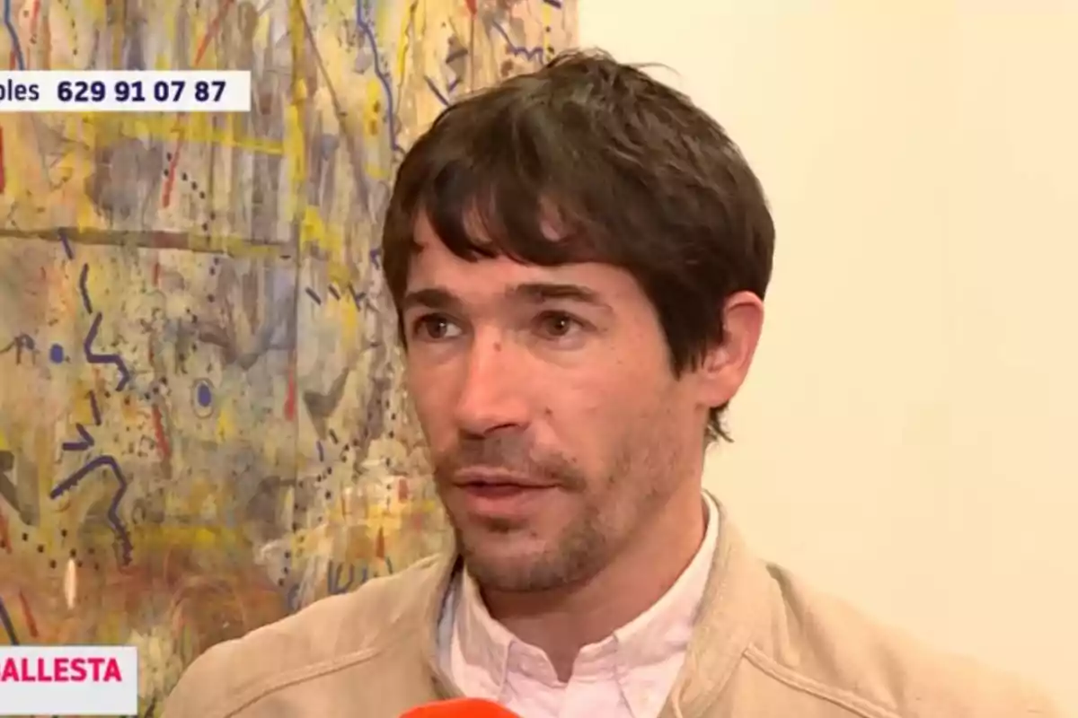 A man with dark hair and a light beard is being interviewed in front of a colorful abstract painting. A man with dark hair and a light beard is being interviewed in front of a colorful abstract painting.