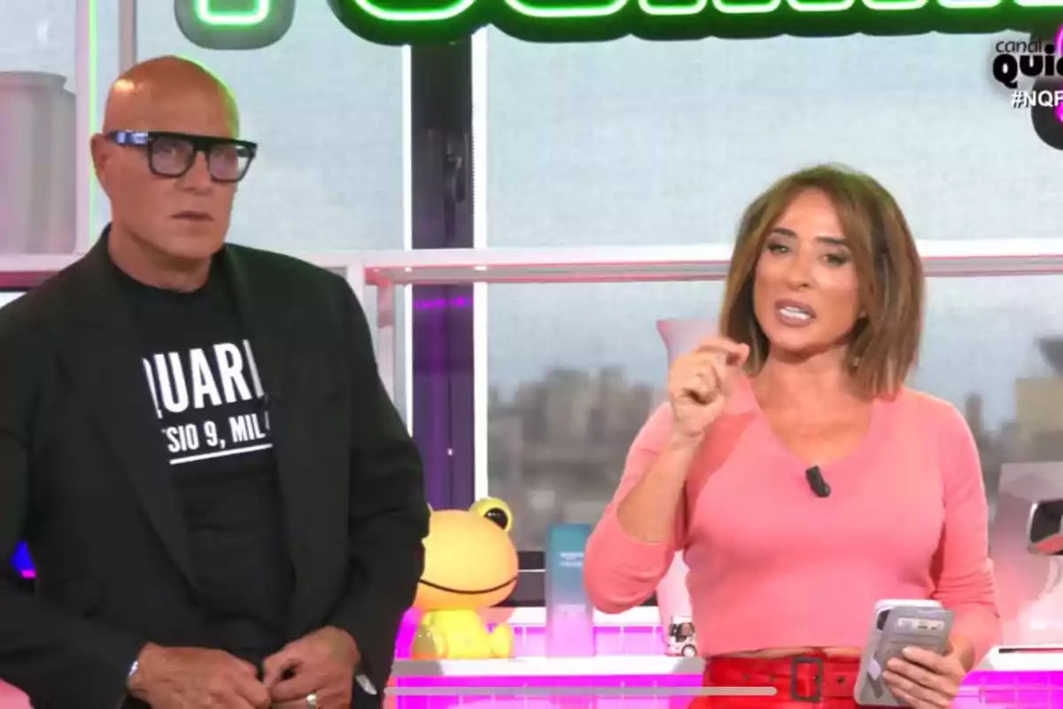 Kiko Matamoros y María Patiño están de pie en un set de televisión, Kiko Matamoros lleva gafas y una camiseta negra, mientras que María Patiño viste una blusa rosa y sostiene un teléfono móvil.