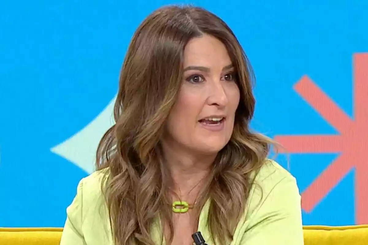 A woman with wavy brown hair speaking on a television show with a blue background and geometric details.