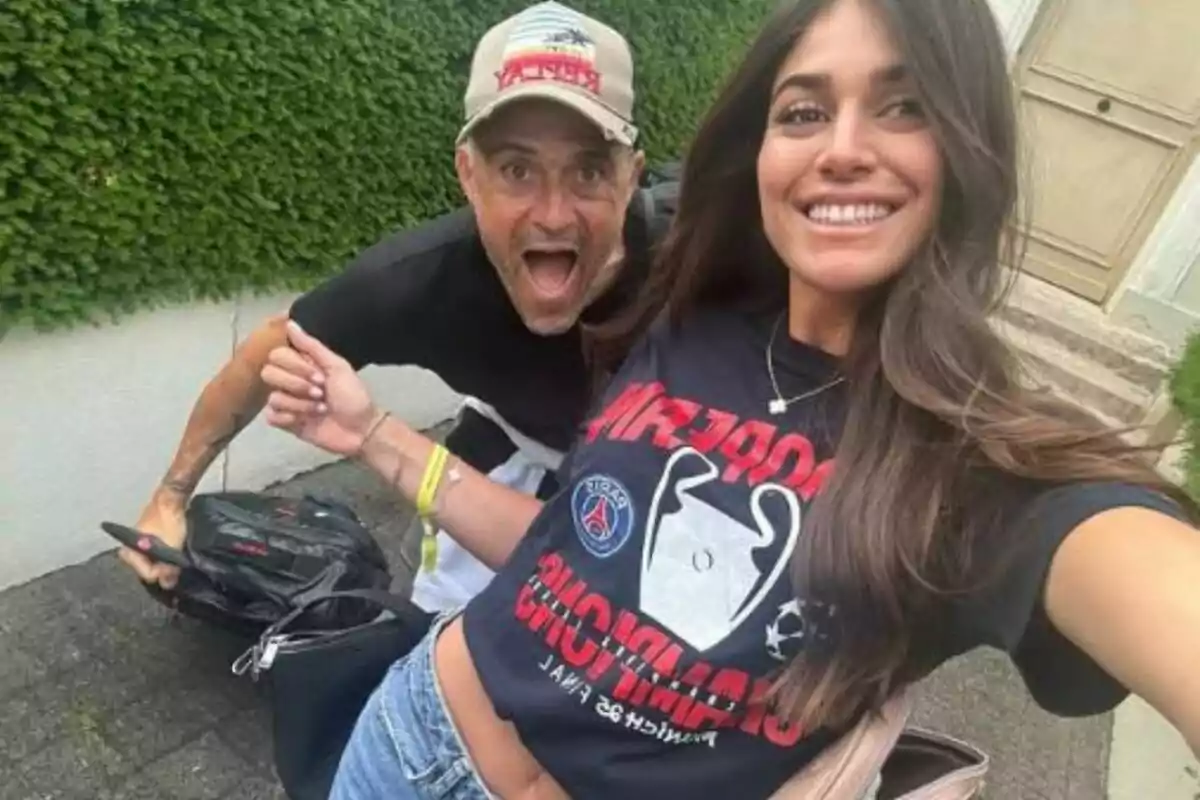 Una jove somrient es fa una selfie al costat d’un home que fa un gest divertit a l’aire lliure davant d’una paret coberta de plantes.