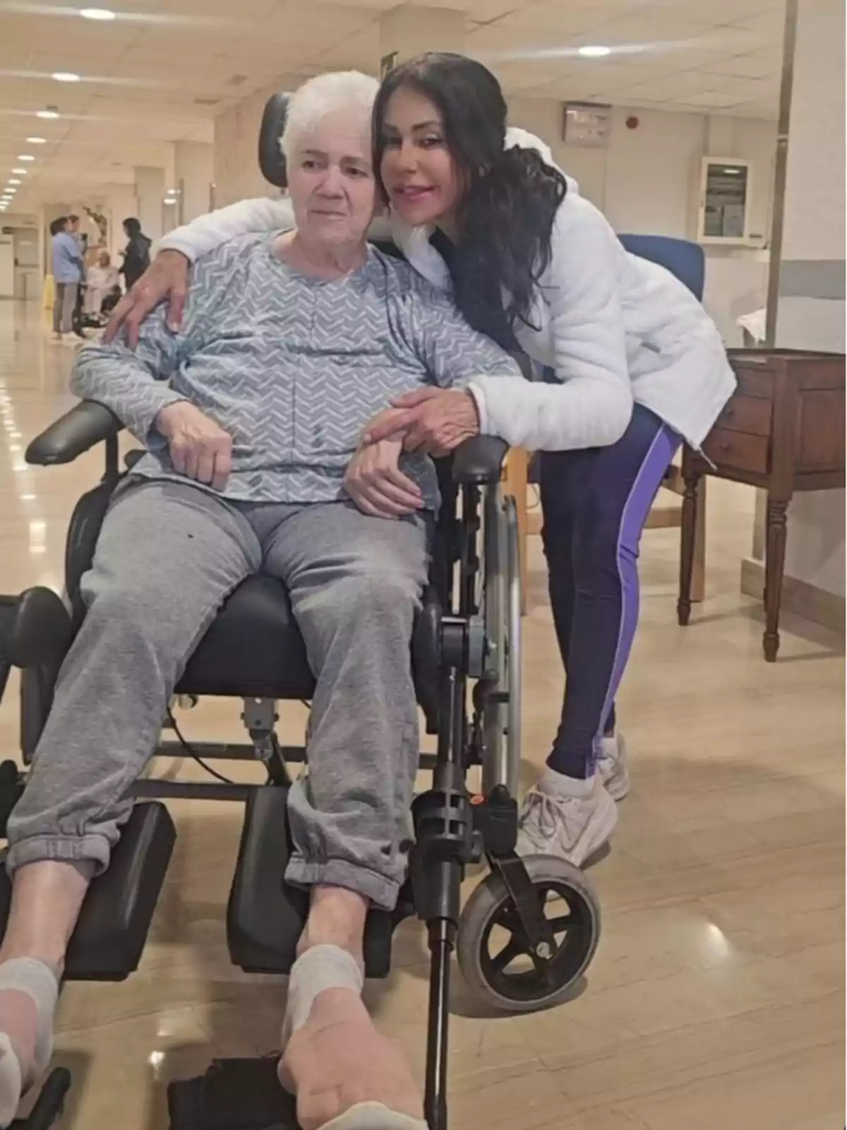 La madre de Maite Galdeano está sentada en una silla de ruedas mientras Maite la abraza y sonríe en un pasillo iluminado.