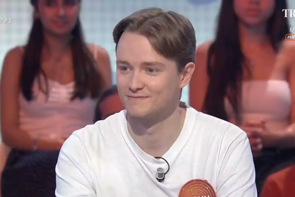 Young person with light-colored hair and a white t-jersey participating in a television show with an audience in the background Young person with light-colored hair and a white t-jersey participating in a television show with an audience in the background