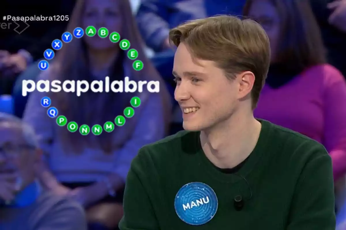 Manu sonríe mientras participa en el programa de televisión "Pasapalabra", con un círculo de letras en el fondo.