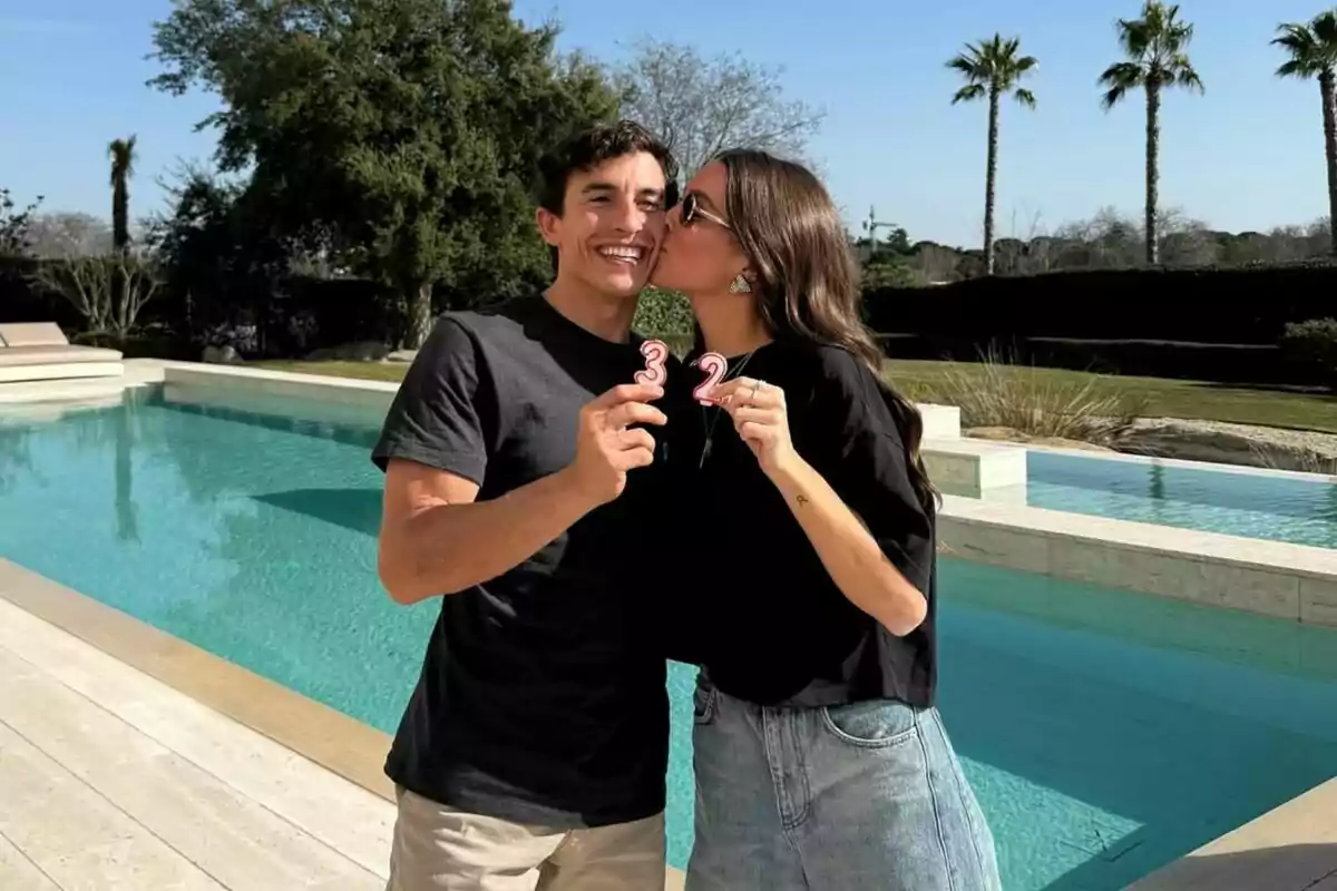 A smiling couple by a pool holds candles with the numbers 3 and 2 while the woman kisses the man on the cheek. A smiling couple by a pool holds candles with the numbers 3 and 2 while the woman kisses the man on the cheek.