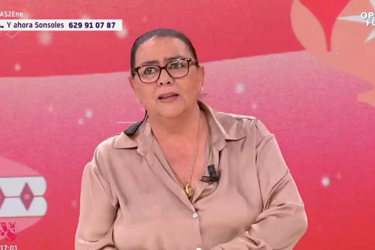 María del Monte con gafas y camisa beige hablando en un programa de televisión con fondo rojo.