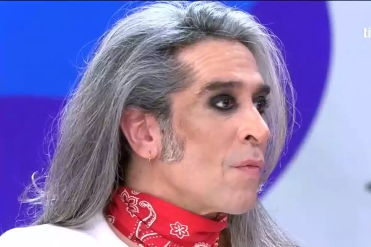 A man with long, gray hair, dark eye makeup, and a red bandana around his neck. A man with long, gray hair, dark eye makeup, and a red bandana around his neck.