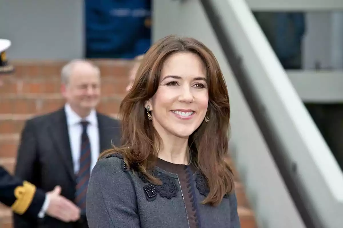 A smiling woman with brown hair and a dark gray coat is standing outdoors with blurred people in the background. A smiling woman with brown hair and a dark gray coat is standing outdoors with blurred people in the background.