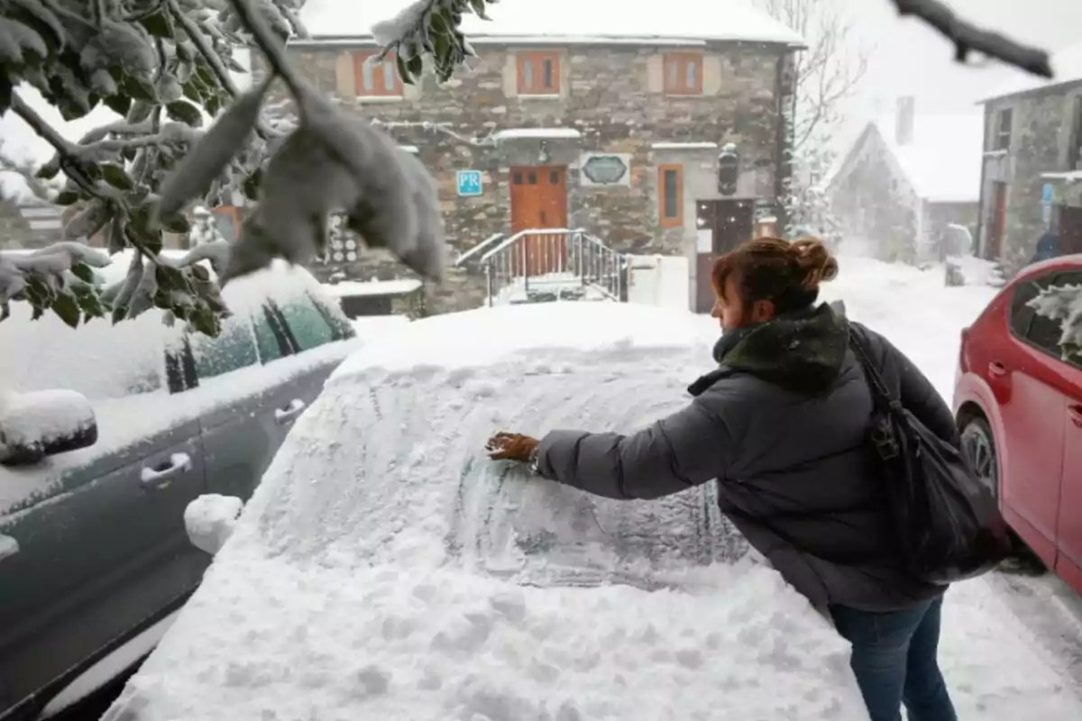 Una persona limpia la nieve acumulada sobre un automóvil en un entorno nevado con edificios de piedra al fondo.