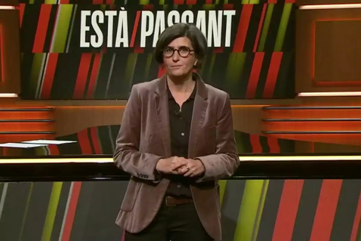Una persona amb ulleres i jaqueta marró està dreta davant d'un fons amb el text Una persona amb ulleres i jaqueta marró està dreta davant d'un fons amb el text