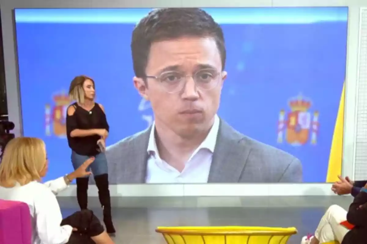 Un programa de televisió amb un gran monitor que mostra un home amb ulleres i vestit mentre diverses persones conversen al set.