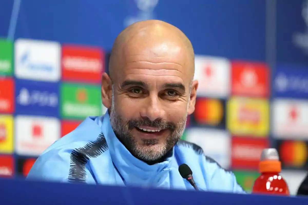 Un hombre calvo con barba sonríe durante una conferencia de prensa con un fondo de logotipos coloridos.