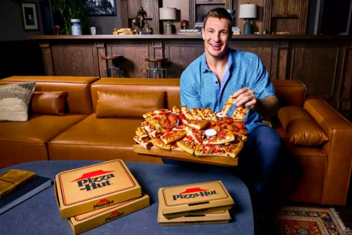 Un home somrient està assegut en un sofà de cuir sostenint una safata plena de diferents tipus de pizza i ales, amb caixes de Pizza Hut sobre la taula davant d'ell en una sala d'estar acollidora. Un home somrient està assegut en un sofà de cuir sostenint una safata plena de diferents tipus de pizza i ales, amb caixes de Pizza Hut sobre la taula davant d'ell en una sala d'estar acollidora.