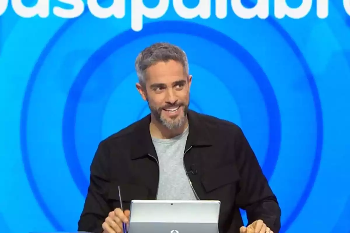 Un home amb barba i cabell canós somriu mentre està assegut davant d'un ordinador portàtil en un plató de televisió amb un fons blau. Un home amb barba i cabell canós somriu mentre està assegut davant d'un ordinador portàtil en un plató de televisió amb un fons blau.