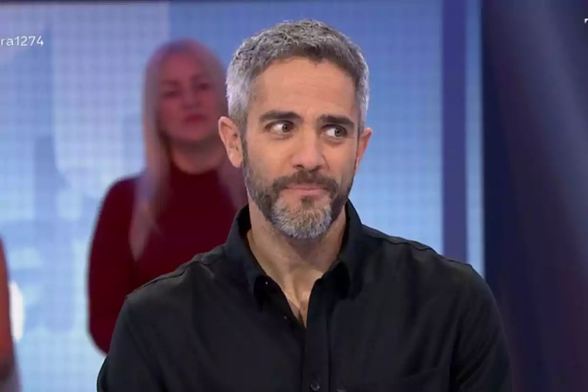 Un home amb barba i cabell canós és en un estudi de televisió amb una expressió pensativa mentre una dona està desenfocada al fons. Un home amb barba i cabell canós és en un estudi de televisió amb una expressió pensativa mentre una dona està desenfocada al fons.
